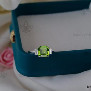 May include: A silver ring featuring a large, emerald-cut, light green gemstone as the central stone, with two smaller, rectangular, white gemstones either side. The ring is presented in a teal jewellery box.