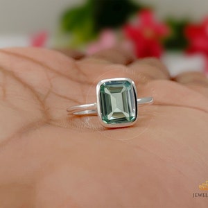 May include: A silver ring with a green emerald-cut gemstone. The ring is on a person's hand.
