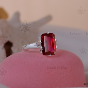 Könnte beinhalten: Ein silberner Ring mit einem großen, rechteckigen, tiefroten Edelstein. Der Ring liegt auf einer rosa Oberfläche, mit den Worten "Jewels of Isha" im Hintergrund. Das Design ist schlicht und konzentriert sich auf die Farbe und Form des Edelsteins.