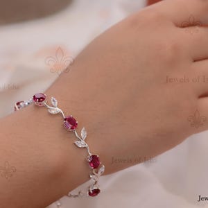 May include: A silver-toned bracelet featuring a series of oval-shaped deep red stones alternating with clear, marquise-cut stones arranged in a vine-like pattern. The metal links connect each stone setting, creating a continuous floral-inspired design.