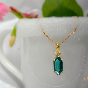 Puede incluir: Un collar de cadena de oro con un colgante de piedra preciosa verde. El colgante tiene forma hexagonal con un corte facetado. El collar está sobre un fondo blanco.