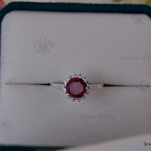 Może przedstawiać: Srebrny pierścionek z dużą, okrągłą, ciemnoczerwoną kamieniem szlachetnym pośrodku, otoczonym mniejszymi, przezroczystymi kamieniami. Pierścionek jest umieszczony w białym pudełku z ciemnozielonym aksamitnym wnętrzem. Pierścionek jest od Jewels of Isha.