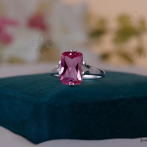 May include: A silver ring with a large, rectangular, pink gemstone. The gemstone is the focal point, set in a simple, elegant band. The ring is displayed on a dark teal surface, with a blurred background.