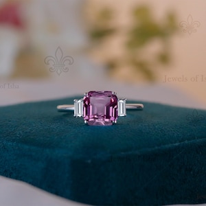 May include: A silver ring with a large, square, purplish-pink gemstone as the centerpiece, flanked by two smaller, rectangular, clear stones. The ring is set on a dark teal velvet surface, with a blurred floral background.