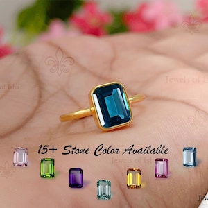 Könnte beinhalten: Ein Goldring mit einem großen, rechteckigen, smaragdgeschliffenen dunkelblauen Edelstein in einer Zargenfassung. Unter dem Ring steht der Text 15+ Stone Color Available, begleitet von acht kleineren rechteckigen Edelsteinen in verschiedenen Farben.