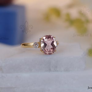 Könnte beinhalten: Ein goldener Ring mit einem großen, oval geschliffenen, hellrosa Edelstein als Mittelpunkt. Zwei kleinere, runde, klare Edelsteine flankieren den Hauptstein. Der Ring liegt auf einer weißen Oberfläche mit unscharfem Hintergrund.