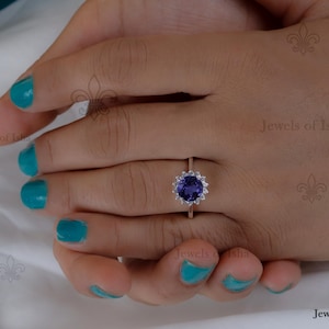 May include: A gold ring featuring a large, round, dark purple gemstone surrounded by smaller, clear stones. The ring is set against a hand with teal nail polish. The ring's design is floral, with the central stone as the focal point.