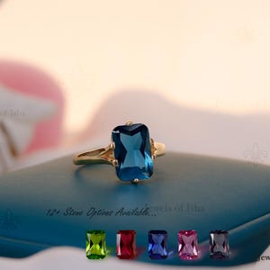 Puede incluir: Un anillo de oro con una gran gema rectangular de color azul oscuro. El anillo está sobre una superficie verde azulado, con el texto "12+ Stone Options Available..." y varias opciones de color de gema debajo. El anillo es una joya.