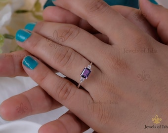 Puede incluir: Un anillo de plata con una piedra rectangular de color púrpura oscuro, flanqueada por dos piedras más pequeñas, redondas y transparentes. El anillo se muestra en una mano con esmalte de uñas turquesa.