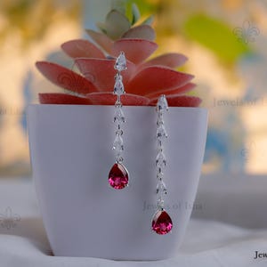 May include: Elegant silver earrings with a dangling design. Each earring features a series of clear, sparkling stones leading to a vibrant, teardrop-shaped, ruby-red gemstone. The earrings are displayed in front of a white pot with a red succulent.