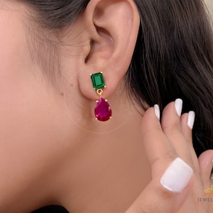 May include: Gold dangle earrings with a green square-shaped gemstone and a teardrop-shaped red gemstone.
