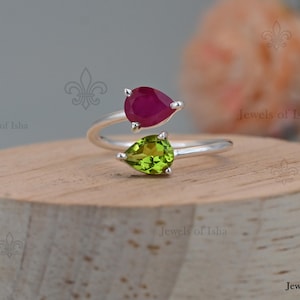 May include: A bypass ring featuring a silver band with two pear-shaped gemstones. One stone is a deep red ruby, and the other is a bright lime green peridot. The ring has an open design, allowing the two stones to sit side by side on the finger.
