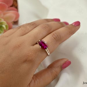 May include: A silver ring featuring a vibrant, rectangular-cut fuchsia gemstone held by four prongs. The band has a split design on the sides, creating a sleek and polished appearance. The ring is worn on a finger, showcasing the bright pink stone.
