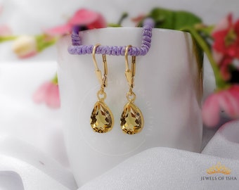 May include: A pair of gold teardrop earrings with yellow gemstones. The earrings are hanging from a white surface with a purple cord in the background.