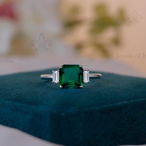 May include: A silver ring featuring a large emerald-cut green gemstone as the central focus, accompanied by two smaller, rectangular white gemstones. The ring is displayed on a dark teal surface, with a blurred floral backdrop.