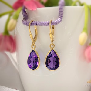 Peut inclure: Une paire de boucles d'oreilles pendantes en or avec des pierres précieuses violettes en forme de goutte. Les boucles d'oreilles sont sur une surface blanche avec des fleurs violettes en arrière-plan.