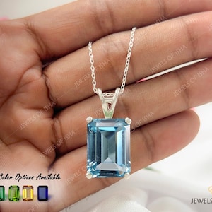 May include: A silver necklace featuring a large, rectangular, emerald-cut blue gemstone pendant held by a four-prong setting. The pendant hangs from a delicate silver chain. Text on the image reads 10+ Color Options Available and JEWELS OF ISHA.