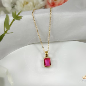 Puede incluir: Un collar de cadena dorada con un colgante cuadrado rosa. El colgante tiene una piedra transparente en el centro.