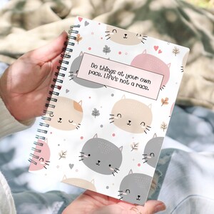 May include: A spiral bound notebook with a white background and a repeating pattern of cartoon cats with pink and gray accents. The notebook has a pink and white sticker with the text "Do things at your own pace. Life's not a race."