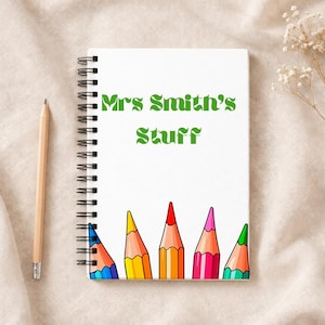 May include: White spiral-bound notebook with the text "Mrs Smith's Stuff" in green. The bottom of the notebook features a row of colourful, cartoon-style pencils. A wooden pencil rests on the left side of the notebook.