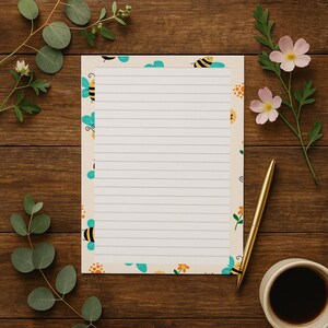 May include: A lined notepad with a bee and floral border. The notepad is cream-colored with a pattern of bees, flowers, and blue cloud shapes. A gold pen and a cup of coffee sit next to the notepad on a wooden surface.