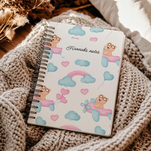 Può includere: Quaderno a spirale con un design stravagante. La copertina presenta un motivo ripetuto di orsetti in aereo, nuvole, cuori e arcobaleni in colori pastello. Il testo "Hannah's notes" è scritto in corsivo.
