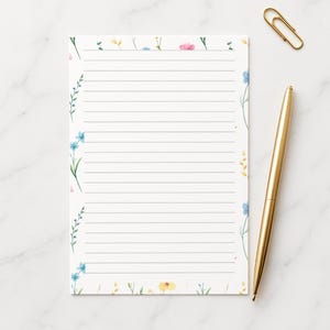 May include: A lined notepad with a floral design. The notepad features delicate watercolor flowers in shades of blue, pink, and yellow along the edges. A gold pen and paperclip are placed next to the notepad on a white marble surface.