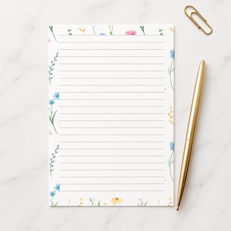 May include: A lined notepad with a floral design. The notepad features delicate watercolor flowers in shades of blue, pink, and yellow along the edges. A gold pen and paperclip are placed next to the notepad on a white marble surface.
