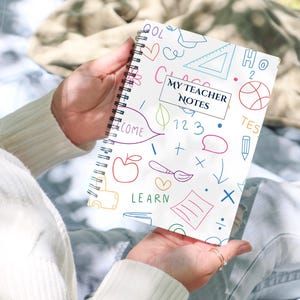 Op de afbeelding: Een wit spiraalgebonden notitieboek met de tekst "MY TEACHER NOTES" op de kaft. De kaft is versierd met kleurrijke doodles van schoolgerelateerde items. Het notitieboek wordt door een persoon vastgehouden.