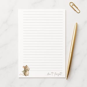 May include: A white rectangular notepad with horizontal lines, featuring a small illustration of a brown mouse wearing a green scarf in the bottom left corner and the handwritten text "don't forget..." in the bottom right, with a gold pen.