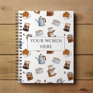May include: A white spiral bound notebook with a coffee themed design. The cover features a repeating pattern of coffee cups, coffee makers, and coffee beans. The text "YOUR WORDS HERE" is printed in the center of the notebook.