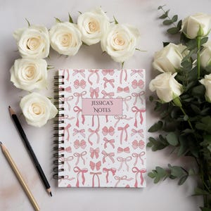 Può includere: Un quaderno a spirale con un motivo a fiocchi rosa e la scritta "JESSICA'S NOTES" su un rettangolo rosa. Rose bianche e matite circondano il quaderno.