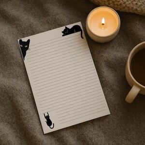 May include: A lined notepad with black cat illustrations in each corner. A lit candle in a white ceramic holder and a mug of coffee sit next to the notepad on a gray surface.