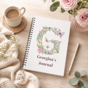 May include: A spiral-bound journal with a white cover featuring a large letter G decorated with watercolor flowers and butterflies. The text Georgina's Journal is printed below in purple. A pen rests beside the journal on a wooden surface.