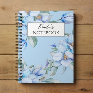 May include: A blue notebook with a floral design featuring white flowers and green leaves. The notebook has a spiral binding and the text "Paula's NOTEBOOK" on the front cover.
