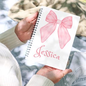 Può includere: Quaderno a spirale con un'illustrazione di fiocco rosa e il nome "Jessica" in corsivo rosa. Il quaderno è aperto, mostrando il disegno su una pagina bianca. Il fiocco è in stile acquerello.