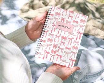 Könnte beinhalten: Ein spiralgebundenes Notizbuch mit weißem Einband und einem wiederkehrenden Muster aus rosa Schleifen in verschiedenen Stilen. Ein rechteckiges rosa Etikett ist mittig auf der Vorderseite platziert, mit dem Wort NOTEBOOK in schwarzen Großbuchstaben.