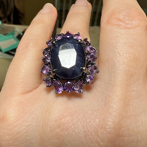 May include: A large, oval-shaped dark blue gemstone ring, surrounded by smaller purple gemstones. The ring has a dark metal setting. The ring is on a person's finger.