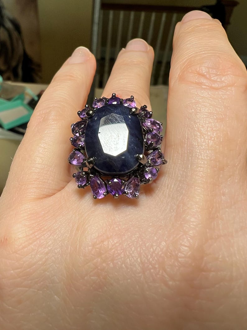 May include: A large, oval-shaped dark blue gemstone ring, surrounded by smaller purple gemstones. The ring has a dark metal setting. The ring is on a person's finger.