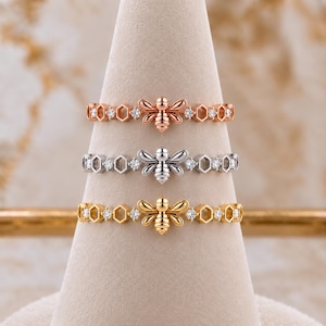 May include: Three rings stacked vertically, each featuring a central bee motif flanked by alternating hexagonal honeycomb shapes and small round clear stones. The rings are finished in rose gold, silver, and yellow gold tones respectively.