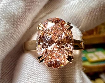 May include: A gold ring featuring a large, oval-cut, peach-colored gemstone. The gemstone is the focal point, with intricate facets catching the light. The ring's band is a simple gold design, and the setting has decorative accents.