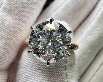 May include: A solitaire ring featuring a large, round, brilliant-cut clear gemstone set in a six-prong metal setting. The band is smooth and polished, showcasing a reflective surface that catches light, creating a bright, sparkling appearance.