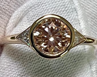 Könnte beinhalten: Ein goldener Ring mit einem großen, runden, champagnerfarbenen Mittelstein in einer glatten Fassung. Zwei kleinere, dreieckig geschliffene, klare Steine sind auf beiden Seiten der zentralen Fassung in die Ringschiene eingesetzt, was ein symmetrisches Design ergibt.