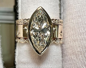 May include: A marquise-cut diamond engagement ring with a gold band. The ring features a large, clear diamond set in a bezel setting. Two additional gold bands with star and dot designs are stacked with the engagement ring. The rings are displayed on a white surface.