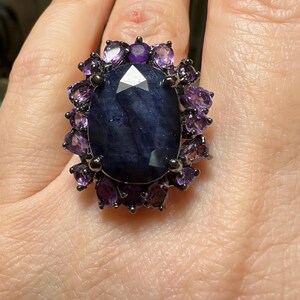 May include: A large, dark blue gemstone ring with a faceted center stone. The ring is surrounded by smaller, purple gemstones and set in a dark metal band. The ring is a statement piece.