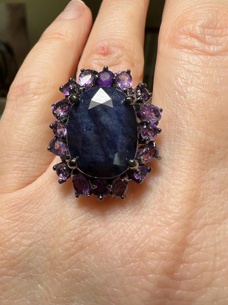 May include: A large, dark blue gemstone ring with a faceted center stone. The ring is surrounded by smaller, purple gemstones and set in a dark metal band. The ring is a statement piece.