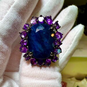 May include: A large, oval-shaped ring featuring a deep blue gemstone at its center, surrounded by smaller, purple gemstones. The ring has a dark metal setting, and the gemstones are set against a white background.