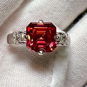 May include: A silver ring featuring a large, faceted, red gemstone as the centerpiece. Two smaller, rectangular, clear gemstones flank the main stone. The ring is set against a white background, highlighting the jewelry's sparkle and design.