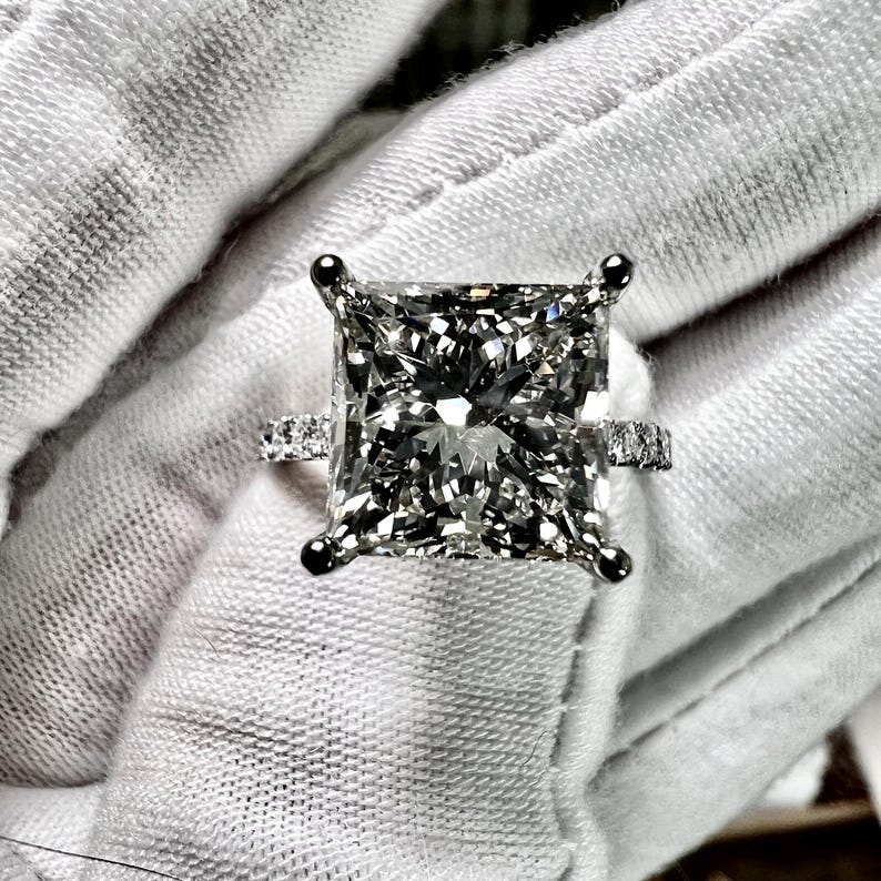 May include: A square-shaped diamond engagement ring with a white gold band and a row of smaller diamonds on each side of the center stone.