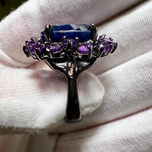 May include: A dark metal ring featuring a large, rectangular, faceted blue gemstone surrounded by smaller, round, purple gemstones. The ring's band is a simple, solid design.